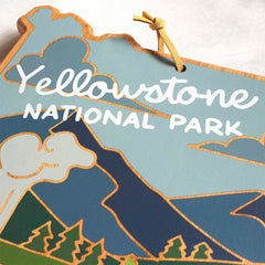 Introducing a decorative wooden sign, crafted to resemble a map and adorned with stylized graphics of mountains, trees, and clouds. This piece features UV printing that displays "Yellowstone National Park" in white text against a light blue background. Its unique design is reminiscent of the Yellowstone Cutting Board with artwork by Summer Stokes from Totally Bamboo.