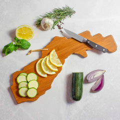 A Grand Canyon cutting board from Totally Bamboo, adorned with artwork by Summer Stokes, showcases a delightful arrangement of sliced lemon, zucchini, a whole zucchini, onion wedges, garlic, rosemary, a knife, and fresh basil on a light gray surface—a true masterpiece.