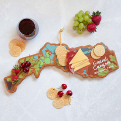 A Grand Canyon Cutting Board with Artwork by Summer Stokes from Totally Bamboo is crafted using a UV printing technique, shaped like the park's outline. It provides an elegant display for cheese, crackers, salami, cherries, strawberries, and grapes. A glass of red wine beautifully complements this stylish presentation.