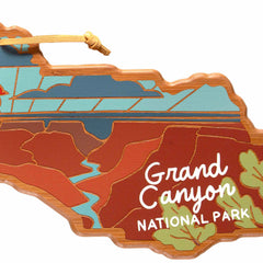 This Grand Canyon Cutting Board by Summer Stokes, presented by Totally Bamboo, is shaped like the park itself and showcases a stylized landscape with a river and canyon beneath a clear blue sky and lush green foliage. Featuring UV printing, the white text elegantly reads "Grand Canyon National Park," resembling an ornament's intricate design.