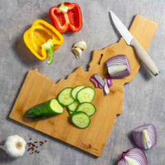 An Idaho Cutting Board with Artwork by Wander on Words™ from Totally Bamboo holds sliced cucumbers, surrounded by red and yellow bell peppers, chopped red onion, garlic, and a knife on a gray surface.