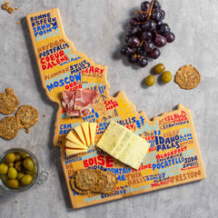 The Idaho Cutting Board with Artwork by Wander on Words™, crafted by Totally Bamboo, showcases vibrant, overlapping city names and is perfect for displaying sliced cheeses and meats. Accompany the spread with grapes and olives, while crackers are stylishly placed on a gray surface.