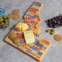 Introducing the Idaho Cutting Board with Artwork by Wander on Words™, crafted by Totally Bamboo. This uniquely styled bamboo cutting board beautifully showcases city names in vibrant text, elegantly shaped like the state of Idaho. It serves as an ideal canvas for an appealing assortment of cheeses and crackers, alongside grapes and a jar of olives. Perfect for those who appreciate state-shaped cutting boards, it is displayed on a sleek gray countertop.