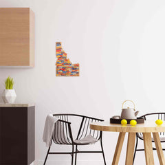 A modern kitchen showcases a sleek black chair and a round wooden table adorned with vibrant yellow lemons. A textured wall map of Idaho embellishes the white wall, complemented by the Idaho Cutting Board by Totally Bamboo featuring artwork from Wander on Words™. Completing the scene is a pristine white teapot and a black cup placed on the table, while a small plant elegantly decorates the counter with its bamboo surface.