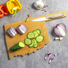 An Oklahoma Cutting Board with Artwork by Wander on Words™ from Totally Bamboo holds a knife, sliced cucumbers, and red onion halves. Nearby are whole and sliced cucumbers, red and yellow bell peppers, a garlic bulb, and scattered peppercorns on the gray surface.