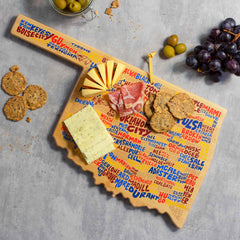 A charcuterie board featuring cheese, cured meats, crackers, and fresh grapes is artfully arranged on a gray surface. The Oklahoma Cutting Board with Artwork by Wander on Words™ from Totally Bamboo is adorned with colorful names of various cheeses and accompanied by a small glass of olives.