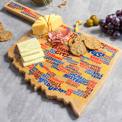 An Oklahoma Cutting Board with Artwork by Wander on Words™, offered by Totally Bamboo, beautifully displays city names while supporting sliced cheese, prosciutto, and crackers. Grapes adorn the scene in the background as a cheese knife rests gracefully on the board's handle—a perfect fusion of functionality and style.