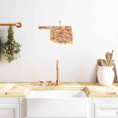 A white kitchen interior with a wooden countertop and copper faucet features an "Oklahoma Cutting Board with Artwork by Wander on Words™" from Totally Bamboo, showcasing colorful typography and hung above the sink. To the right, additional state-shaped cutting boards complement the wooden utensils and pot.