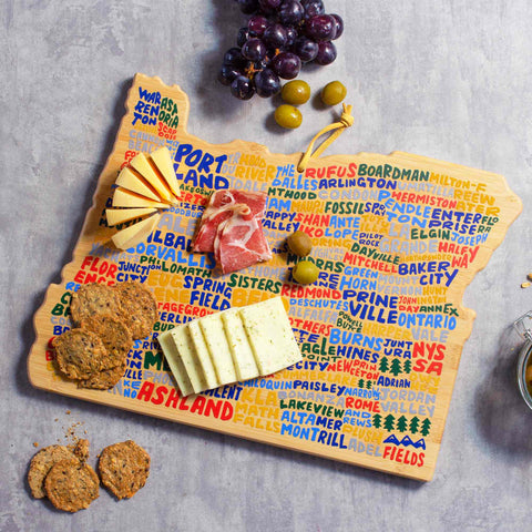 The Oregon Cutting Board with Artwork by Wander on Words™, created by Totally Bamboo, is a wooden cutting board featuring brightly illustrated city names in hand-lettered style. This charming setup includes an assortment of cheeses, crackers, cured meats, olives, and grapes artfully presented on a stone surface with additional crackers served alongside.