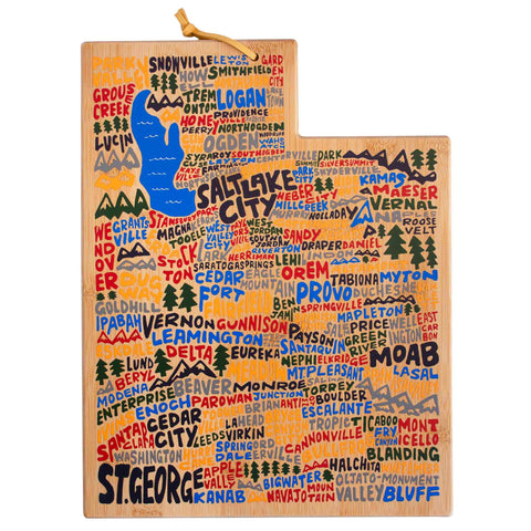 The Utah Cutting Board with Artwork by Wander on Words™, crafted by Totally Bamboo, is designed in the shape of Utah and adorned with vibrant, hand-painted and hand-lettered artwork. It highlights towns and cities such as Salt Lake City, St. George, and Logan, along with illustrations of a lake and mountains.