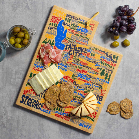 The Utah Cutting Board with Artwork by Wander on Words™, crafted by Totally Bamboo, holds sliced meats, cheese, and crackers. A small glass dish contains olives, and a bunch of grapes rests on the gray surface nearby.